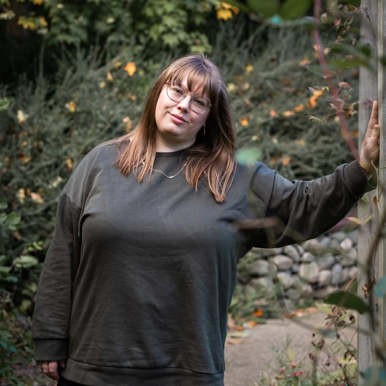 A person with long brown hair stands in a garden, leaning against a wall.