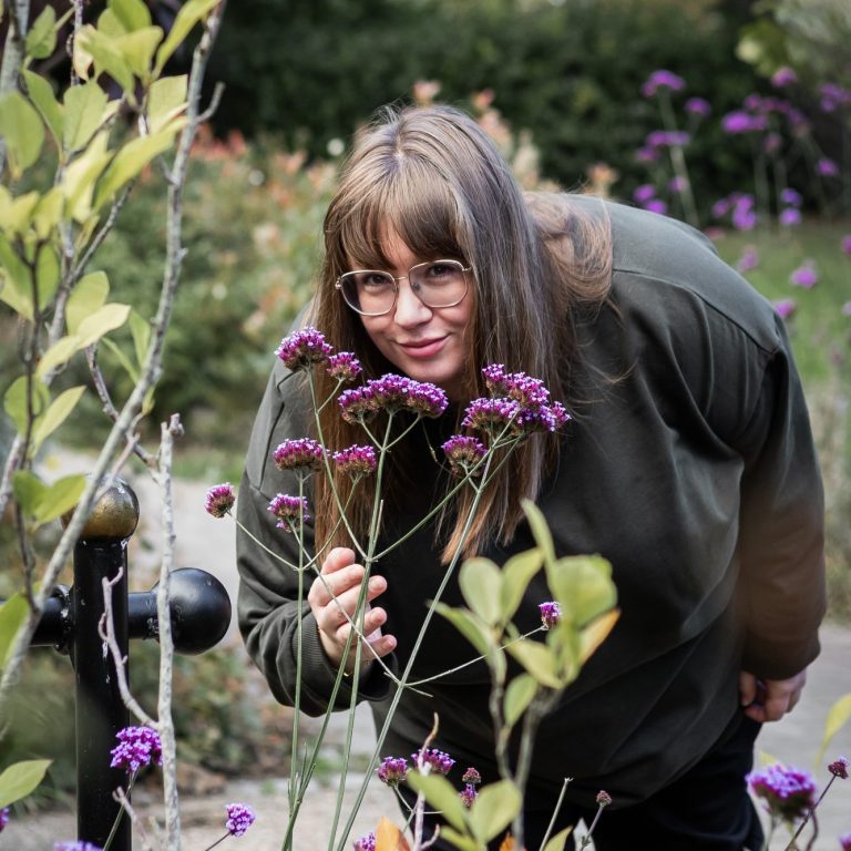 Rachel Conductive Education specialist in motor disorders A person with glasses leans forward to smell flowers in a garden setting.